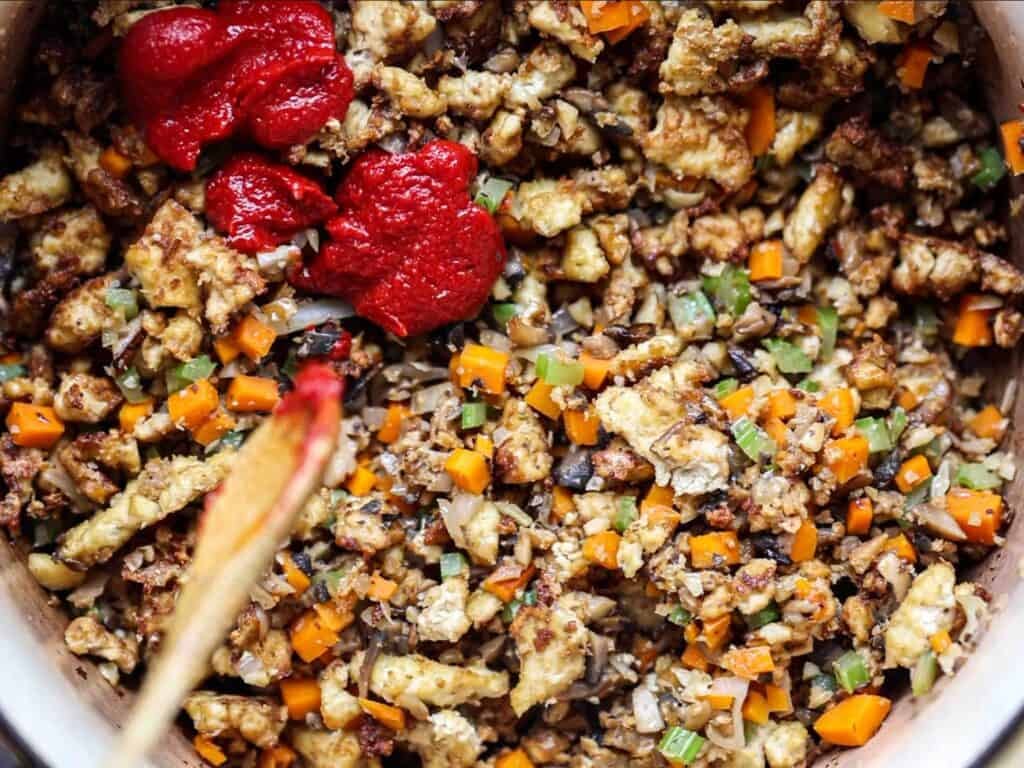 Close-up of a pan filled with sautéed chopped vegetables, browned crumbled tofu, and two dollops of tomato paste, being stirred with a wooden spoon.
